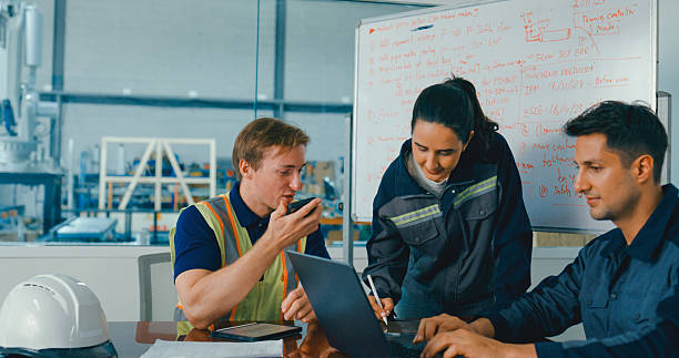 Three industrial workers collaborating at a workstation, reviewing data on a laptop in a factory setting.