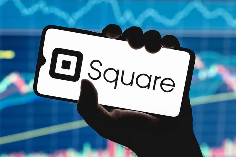 Hand holding a smartphone displaying the Square logo against a market chart background.
