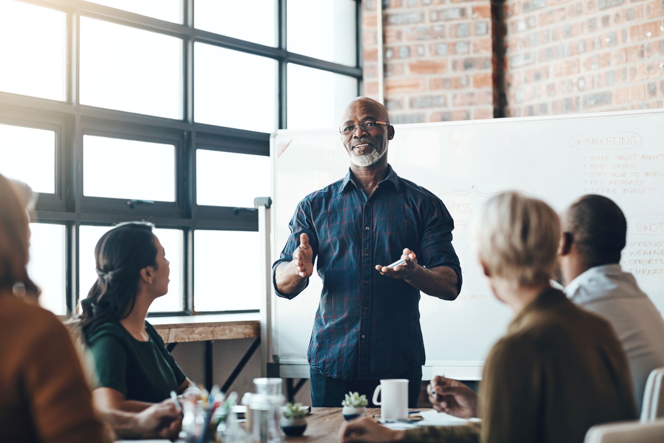 Man leading a team meeting or workshop in front of a whiteboard, engaging a group of diverse professionals.