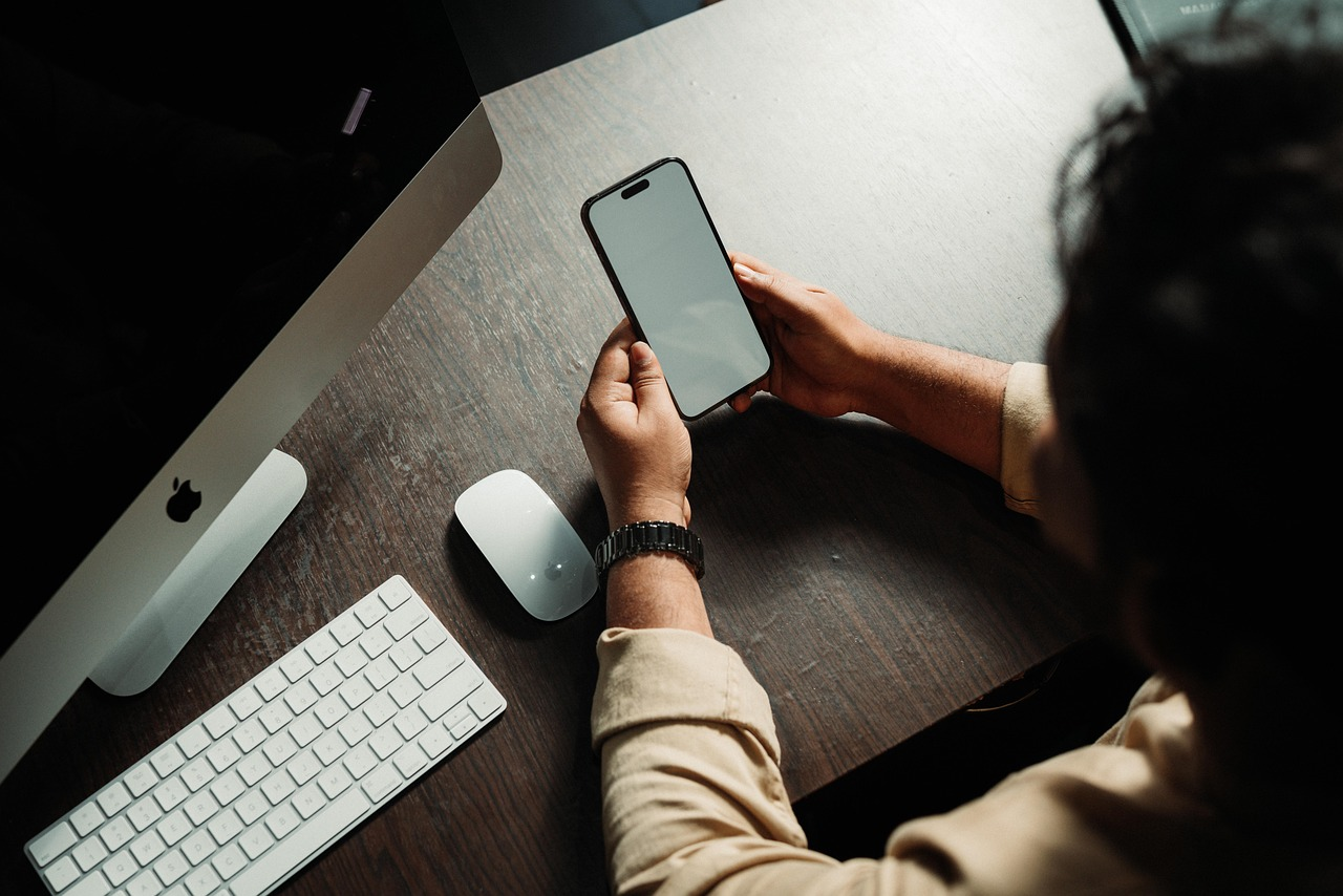 Person sitting at a desk holding a smartphone with a blank screen, next to an Apple computer and keyboard.