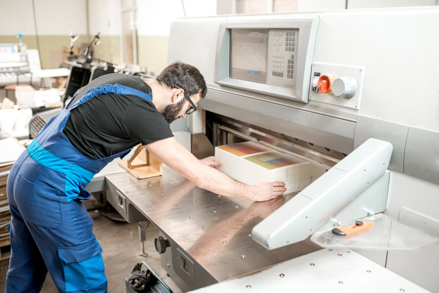 Worker operating a large industrial printing machine with a stack of printed sheets.