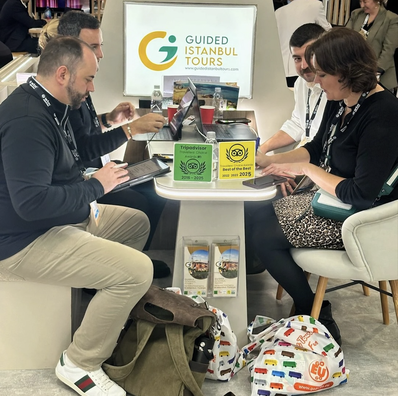 Four people sit around a table at a Guided Istanbul Tours booth during a travel expo, working on laptops beside Tripadvisor award plaques.
