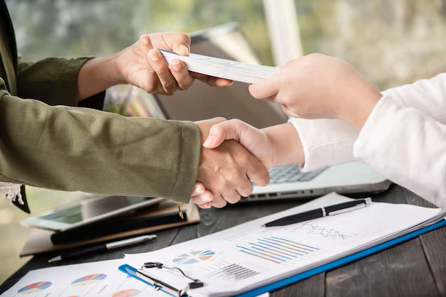 Two people shaking hands across a desk with paperwork and a laptop.